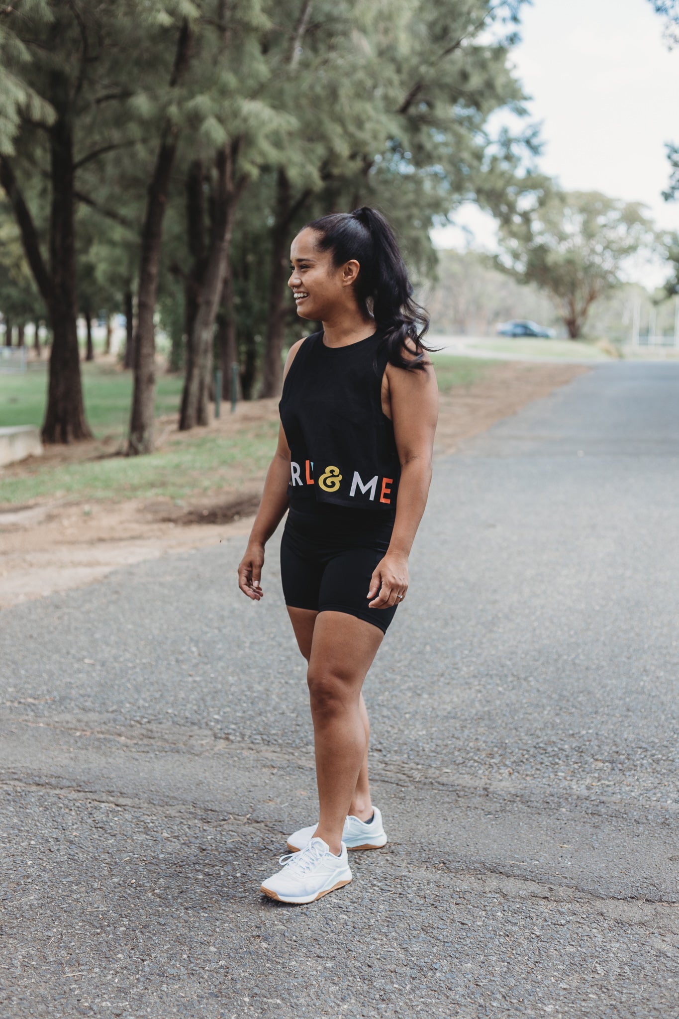 Model wearing the Classic Black Crop Tank Top with the Marlz and Me Logo banding the bottom of the crop. Also wearing the Classic Black Bike Shorts while taking a walk at a sporting field on a sunny day.