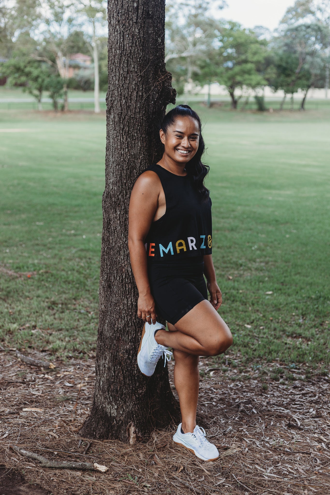 Model wearing the Classic Black Crop Tank Top with the Marlz and Me Logo banding the bottom of the crop. Also wearing the Classic Black Bike Shorts while at a sporting field on a sunny day.