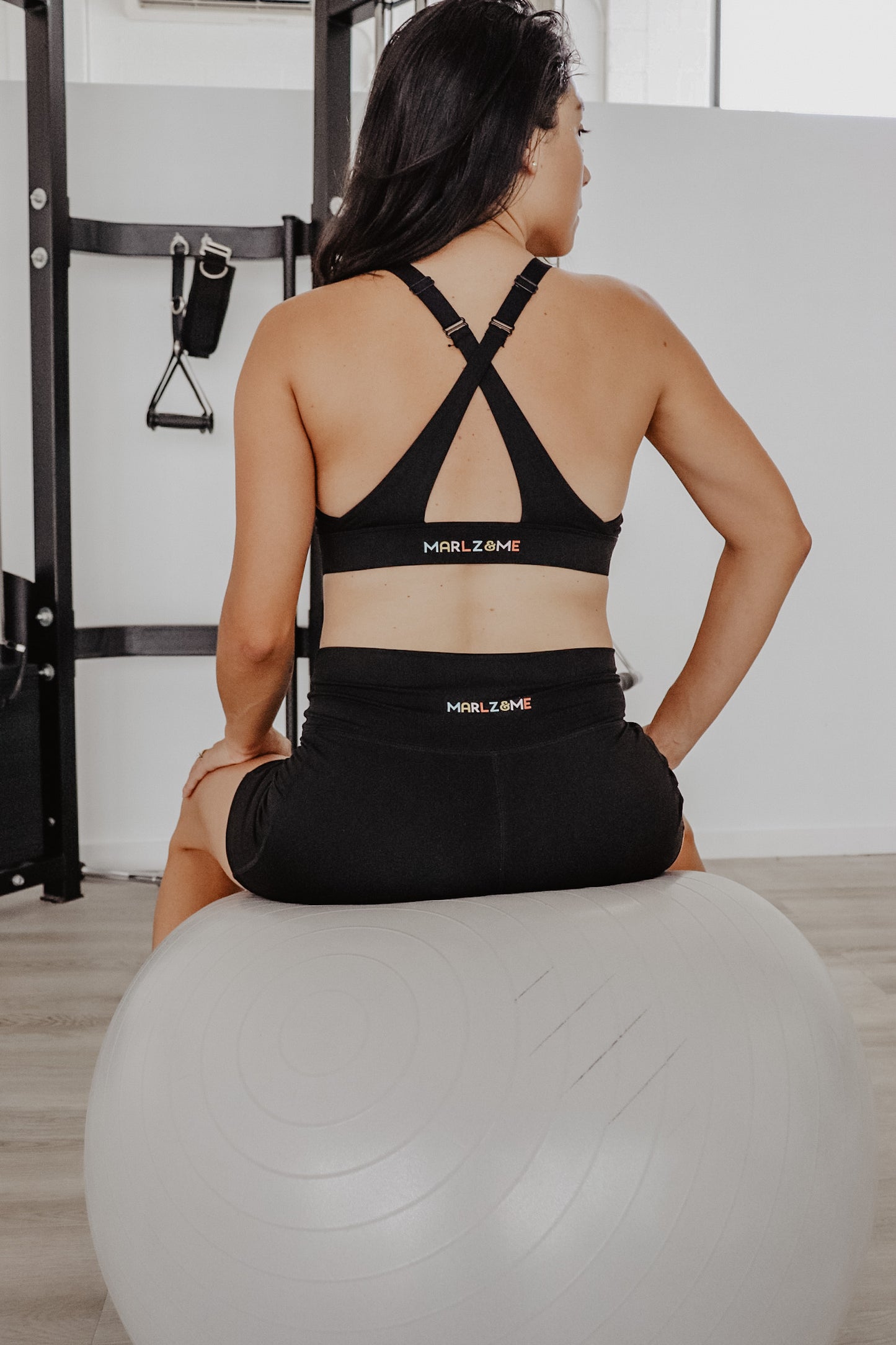 Women at the gym wearing the Classic Black Cross Sports Bra and Classic Black Bike Shorts. View from the back showing the Marlz and Me Logos