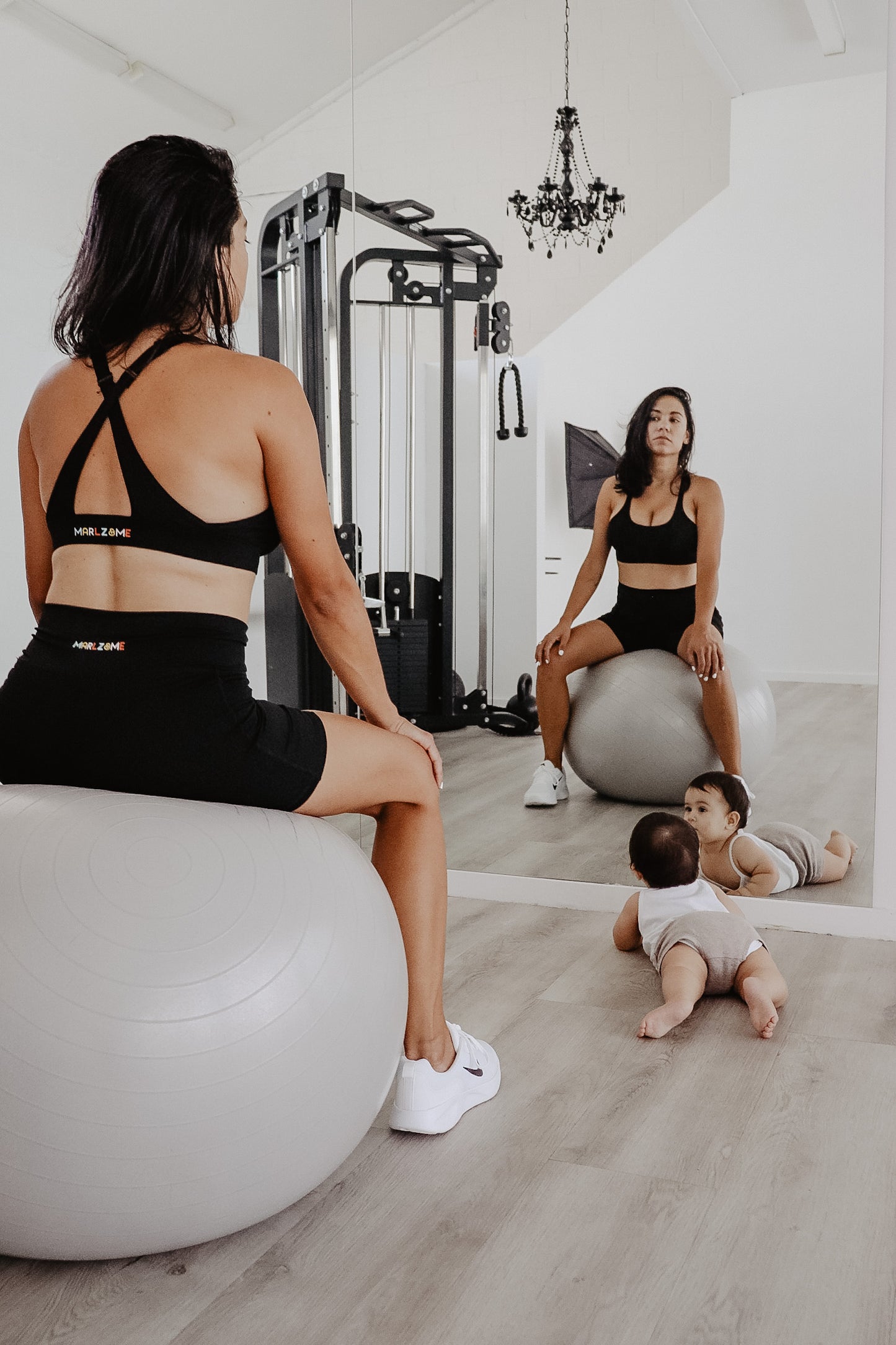 Model wearing Black Sports Bra and Bike Shorts while exercising in private gym using a ball while her baby looks on.